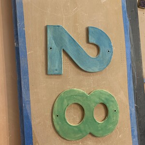 May include: A blue ceramic number "2" and a green ceramic number "8" are displayed on a light brown surface. Each number has mounting holes. The background features a blue-painted wooden frame.
