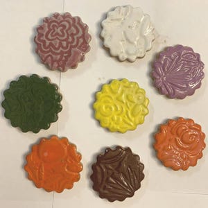 Puede incluir: Ocho posavasos de cerámica con diferentes diseños. Los posavasos son redondos y tienen un borde festoneado. Los diseños incluyen un patrón floral, un patrón geométrico y un color sólido. Los posavasos son de varios colores, incluyendo morado, verde, amarillo, naranja, marrón y blanco.