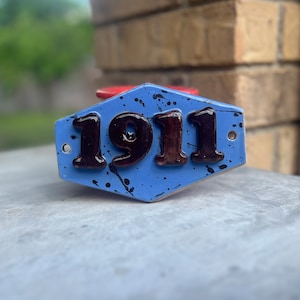 May include: A blue ceramic sign with black paint splatters and the number 1911 in raised black lettering.