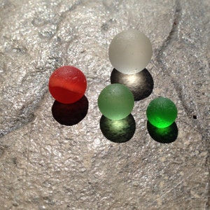 May include: Four glass marbles in different colors: red, white, green, and green. The marbles are arranged in a triangle shape on a gray textured surface.