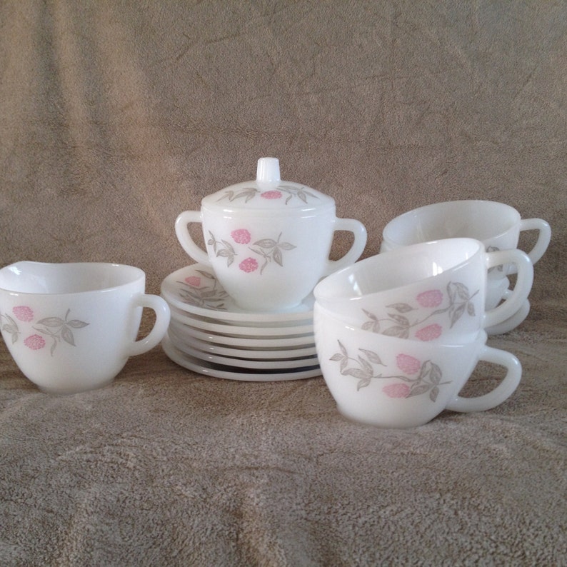 May include: A set of white milk glass teacups and saucers with a pink and grey floral design. The set includes a creamer and sugar bowl with lids.