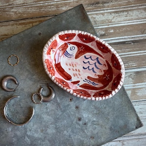 May include: An oval ceramic trinket dish featuring a hand-painted pink and red bird motif with scalloped edges, accompanied by a collection of metal jewelry including a thin gold ring, two silver hoop earrings, and a silver cuff bracelet.