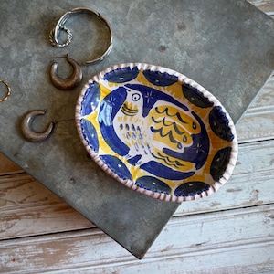 May include: An oval ceramic trinket dish featuring a hand-painted yellow and blue bird motif with a scalloped edge, accompanied by a silver-toned twisted bangle, two small hoop earrings, and a thin gold-toned ring.
