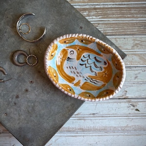 May include: An oval ceramic dish featuring a hand-painted bird motif in shades of ochre, blue, and white, surrounded by three metal hoop earrings and a thin gold-toned ring. The dish has a scalloped edge and a glossy finish.
