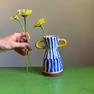 May include: A ceramic vase with a white base and vertical blue stripes, featuring two yellow handles on the sides and a reddish-brown base. The vase has a flared neck and a tapered body, with one yellow flower placed inside the opening.