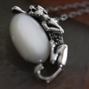 Puede incluir: Un collar con colgante plateado que presenta una figura estilizada abrazando una gran piedra ovalada blanca. La figura está detallada con pequeños acentos oscuros y está unida a una delicada cadena. El colgante tiene una estética vintage o antigua.