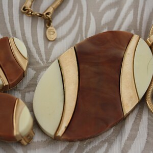 May include: A gold chain necklace with a large oval pendant. The pendant is made of brown and white plastic with gold accents. The pendant has a geometric design. The necklace comes with a pair of matching earrings.