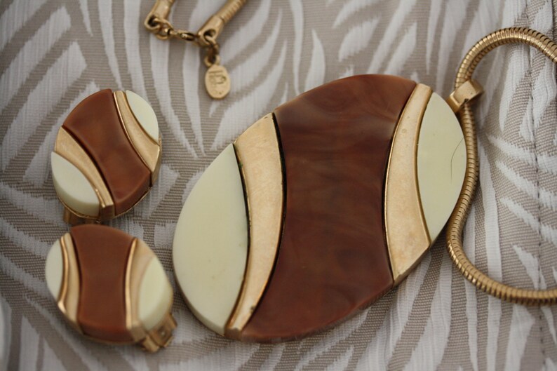 May include: A gold chain necklace with a large oval pendant. The pendant is made of brown and white plastic with gold accents. The pendant has a geometric design. The necklace comes with a pair of matching earrings.