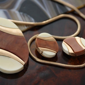May include: A gold chain necklace with a brown and white oval pendant and matching earrings. The pendant and earrings have a geometric design with gold accents.
