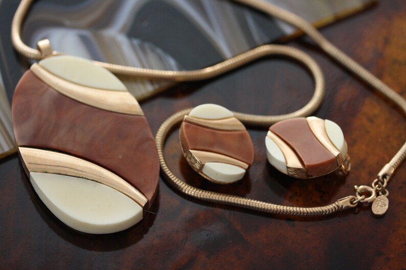 May include: A gold chain necklace with a brown and white oval pendant and matching earrings. The pendant and earrings have a geometric design with gold accents.