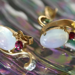 May include: A pair of gold-toned earrings with oval, iridescent white stones and small, round, red gemstones. The earrings have a curved design with a small cluster of clear stones at the end of the curve.