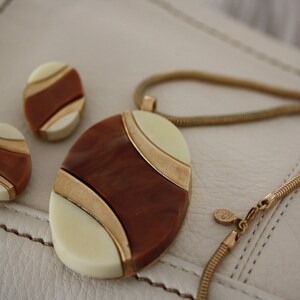 May include: A gold chain necklace with a brown, white, and gold oval pendant. The pendant has a geometric design. The necklace comes with a pair of matching earrings.