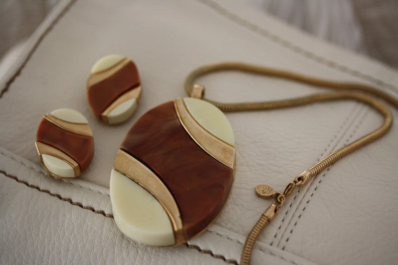 May include: A gold chain necklace with a brown, white, and gold oval pendant. The pendant has a geometric design. The necklace comes with a pair of matching earrings.