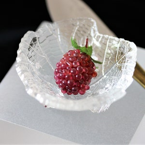 Könnte beinhalten: Ein einzelnes, rotes, beerenförmiges Objekt aus kleinen, runden Perlen sitzt in einer durchsichtigen Glasschale mit einem blattartigen Design. Die Schale steht auf einer weißen Oberfläche.