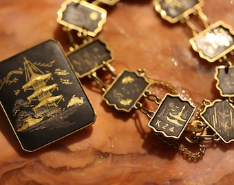 Op de afbeelding: Een zwarte en gouden ketting met een ketting en meerdere rechthoekige hangers. Elke hanger heeft een gouden ontwerp op een zwarte achtergrond. Een hanger heeft de tekst "K24" erop.