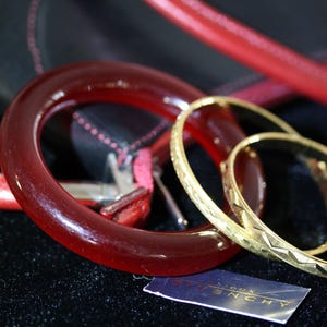 Puede incluir: Un conjunto de tres pulseras, una roja y dos doradas. La pulsera roja está hecha de un material liso y brillante, mientras que las pulseras doradas están hechas de un metal más texturizado. Las pulseras están dispuestas sobre una superficie negra.