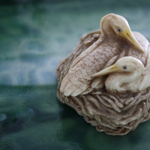 May include: A detailed sculpture of two birds nestled in a nest. The birds have cream-colored bodies, yellow beaks, and blue eyes. The nest is a textured brown, set against a blurred green background. The sculpture is likely a decorative item.