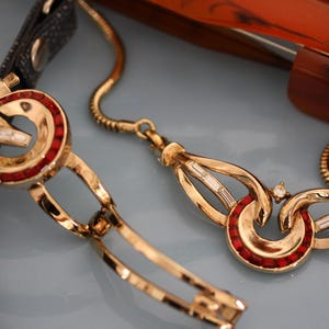 Op de afbeelding: Close-up van een goudkleurige ketting met rode en heldere accenten. De ketting heeft een uniek ontwerp met gebogen schakels en een decoratieve hanger. Een zwarte leren band en een bruin object zijn op de achtergrond.