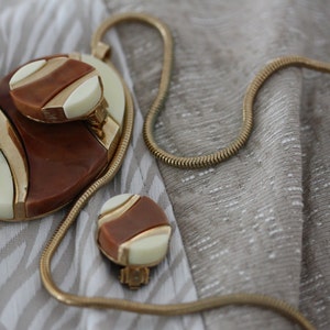 May include: A gold chain necklace with a brown and white oval pendant and matching earrings. The pendant has a geometric design with gold accents.