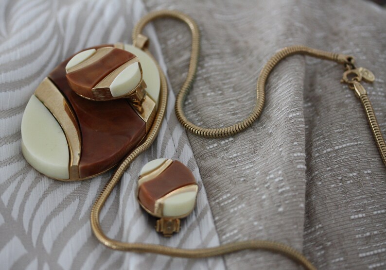 May include: A gold chain necklace with a brown and white oval pendant and matching earrings. The pendant has a geometric design with gold accents.