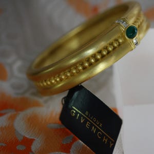 Puede incluir: Un brazalete dorado con una piedra verde y pequeñas piedras transparentes. El brazalete tiene un diseño texturizado con una fila de pequeñas cuentas. Una etiqueta negra con el logo de Givenchy Bijoux es visible.
