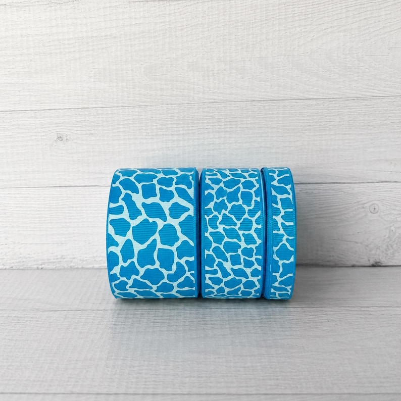 Puede incluir: Tres carretes de cinta de grosgrain con un patr&oacute;n de jirafa en azul brillante y blanco. Las cintas est&aacute;n dispuestas horizontalmente, mostrando la superficie texturizada de la tela y el dise&ntilde;o repetitivo de manchas animales en cada carrete.