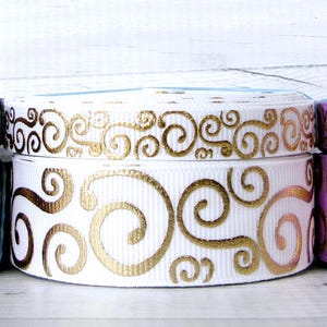 May include: Three spools of grosgrain ribbon featuring a metallic gold swirl pattern. The ribbons are in deep purple, white, and light lavender. Each spool is wound into a circular shape, displaying the repeating gold design on the textured fabric.
