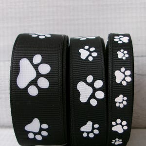 May include: Three rolls of black ribbon featuring white paw prints. The ribbon widths vary, with the paw prints clearly visible. The background is a light, textured surface.