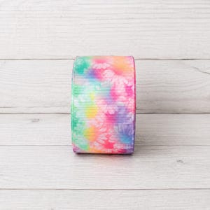 May include: A spool of grosgrain ribbon featuring a vibrant tie-dye pattern with blended shades of pink, yellow, green, blue, and purple. The ribbon has a distinct horizontal ribbed texture and is printed with a repeating white daisy flower motif.