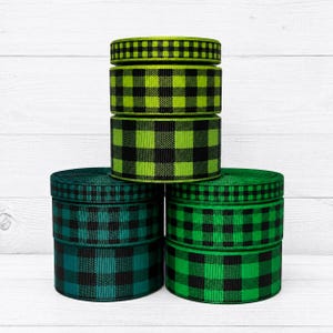 May include: A stack of five spools of ribbon featuring a black and green buffalo plaid pattern. The ribbons are arranged in a pyramid formation, showcasing varying shades of green including lime and forest green, all with a consistent checkered design.