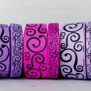 May include: Six rolls of decorative ribbon in shades of purple and pink, each featuring a black swirl design. The ribbons are arranged side by side against a white wooden background, showcasing their contrasting colors and patterns.