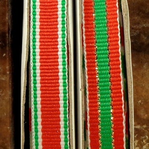 May include: Two rolls of ribbon with a woven design. One ribbon features red, white, and green vertical stripes. The other ribbon has a red and green striped pattern with a gold metallic edge. The ribbons are in a cardboard container.