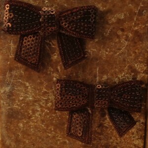May include: Two brown sequined bows with a brown velvet-like texture. The bows are on a brown textured background.