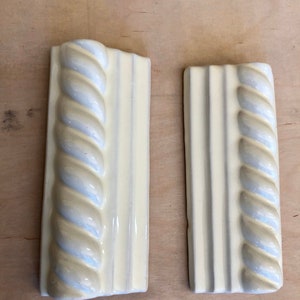 May include: Two white ceramic tile trim pieces with a rope design. The tile on the left has a rounded edge. The tile on the right has a straight edge.