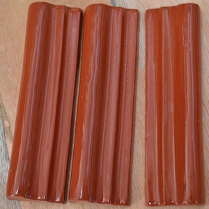 May include: Three terracotta-colored ceramic tiles with a wavy, ribbed design. The tiles are arranged horizontally and have a glossy finish.