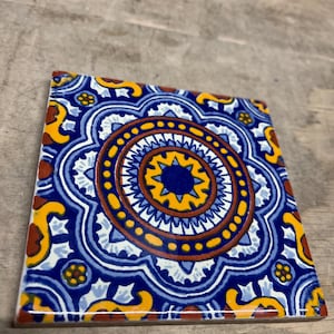May include: A square ceramic tile with a colorful, intricate design. The tile features a blue, yellow, and red pattern with a central circle surrounded by concentric rings.