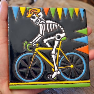 May include: A colorful ceramic tile with a black background features a skeleton riding a yellow bicycle with blue and black wheels. The skeleton is wearing a yellow helmet and has a green leaf on its handlebars. The tile is decorated with colorful triangles around the border.