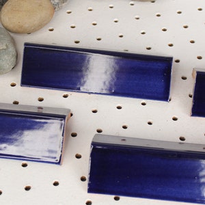 May include: Four blue ceramic tiles with a white glaze. The tiles are arranged in a row on a white pegboard background.