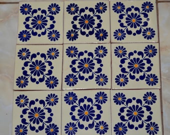 Puede incluir: Nueve baldosas de cerámica cuadradas con fondo color crema. Cada baldosa presenta un diseño floral azul con un centro naranja. El patrón floral es simétrico, con cuatro tallos curvos y flores más pequeñas en cada esquina. Las baldosas están dispuestas en una cuadrícula de 3x3.