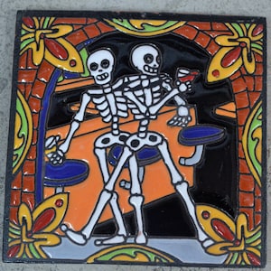 Puede incluir: Dos figuras de esqueleto, pintadas de blanco, se representan en un diseño colorido de tipo azulejo. Están sentados en un bar, uno de ellos sosteniendo un vaso de líquido rojo. El fondo es una mezcla vibrante de naranja, verde y azul.