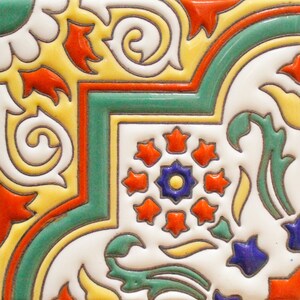 May include: A close-up of a colorful ceramic tile with a geometric pattern. The tile features a central motif of a blue and yellow flower surrounded by red stars. The tile is decorated with swirls and leaves in shades of green, yellow, and orange.