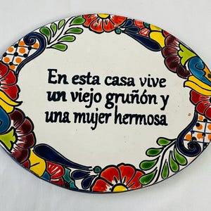 Könnte beinhalten: Ein weißer ovaler Keramikteller mit einem bunten floralen Rand. Der Teller trägt einen spanischen Spruch in schwarzer Schrift: "En esta casa vive un viejo gruñón y una mujer hermosa".