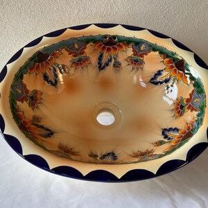 Pode incluir: Uma pia de banheiro de cerâmica com uma borda azul e branca festonada. A pia é decorada com um padrão floral colorido com girassóis e outras flores.