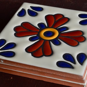 May include: A stack of square ceramic tiles with a floral design. The flower has red, blue, and yellow accents on a white background. The tiles are stacked on top of each other.