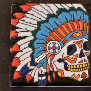 May include: A ceramic tile with a colorful illustration of a skull wearing a headdress with feathers. The skull is white with black outlines and features a colorful geometric design. The headdress is blue, green, red, and orange.