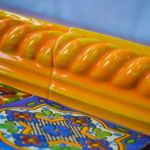 May include: Close-up of a yellow and orange ceramic tile with a rope-like design. The tile is part of a larger pattern of colorful tiles with floral designs.