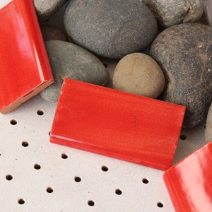 May include: Four bright red rectangular ceramic tiles with rounded edges, arranged on a white surface with small holes. The tiles are surrounded by smooth, gray and brown stones.