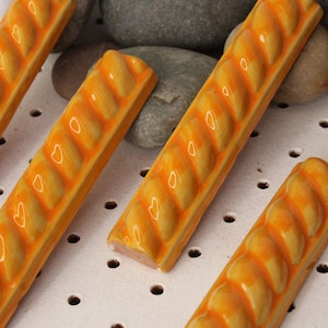 May include: Four orange ceramic tiles with a rope-like design. The tiles are arranged in a row on a white surface with small holes.