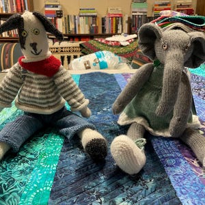 May include: Two handmade knitted dolls: a dog and an elephant. The dog doll has a black and white head, a striped sweater, blue jeans, and a red scarf. The elephant doll is gray with a green dress and white shoes. They are sitting on a colorful quilt.