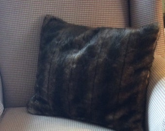 May include: A dark brown faux fur pillow rests on a patterned armchair. The pillow has a rectangular shape and a plush texture, providing a cozy aesthetic. The chair's fabric features a small, checkered pattern in neutral tones, complementing the pillow's rich color.